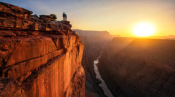 the sun rests over the horizon as people look on from the edge of the grand canyon