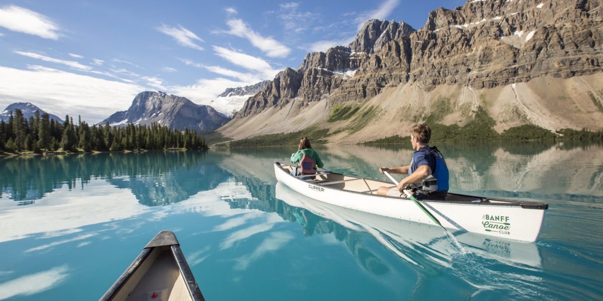people in canoes glide across blue water with tall mountains in background