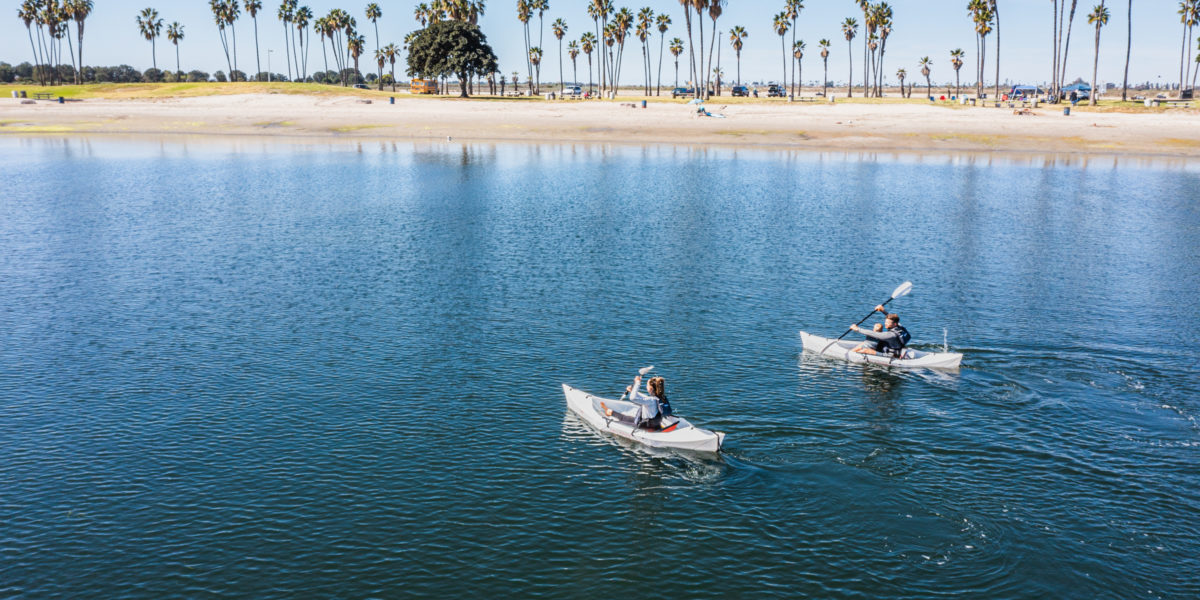 two people in kayaks on smooth water, beach and palm trees in distance