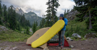 woman blows into sleeping mat to inflate it with a tent, trees, and mountains in background