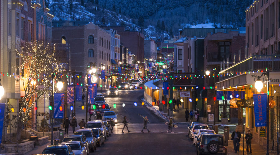 A family of ski and snowboard enthusiasts cross a festively lit and decorated Main Street Park City.