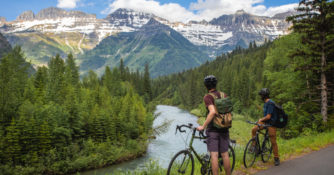 two cyclists stand beside bikes looking out at a river and mountains
