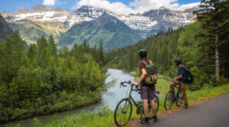 two cyclists stand beside bikes looking out at a river and mountains