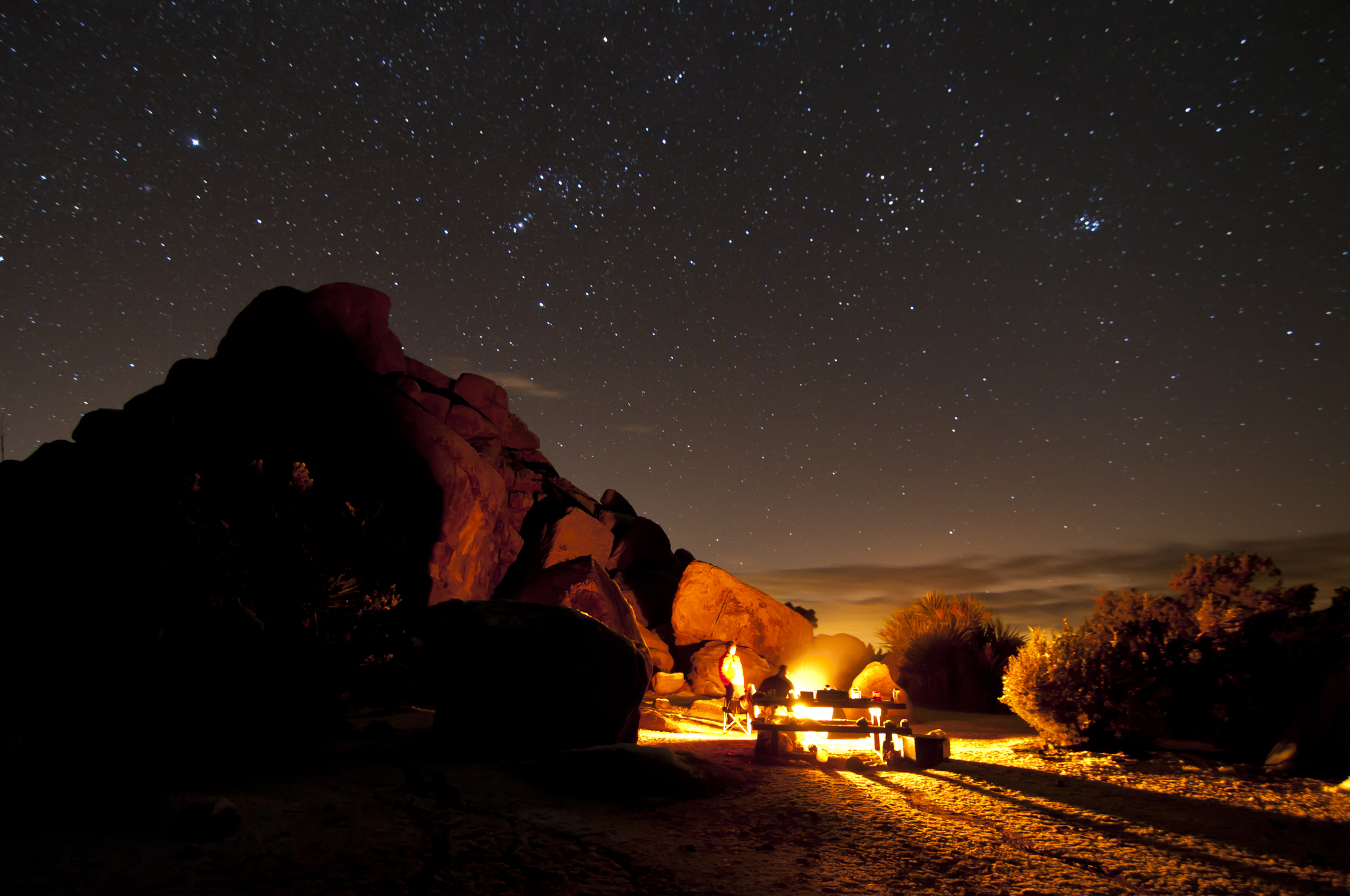People camping next to a boulder with campfire at Joshua Tree at night with stars overhead
