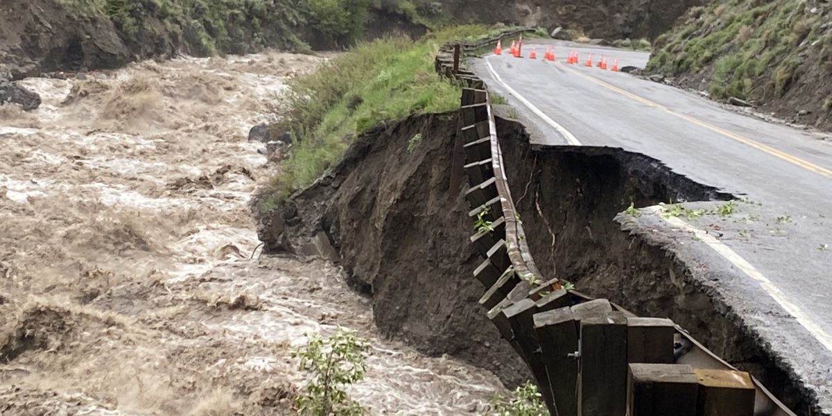 river water rushes next to a collapsed portion of road after flooding